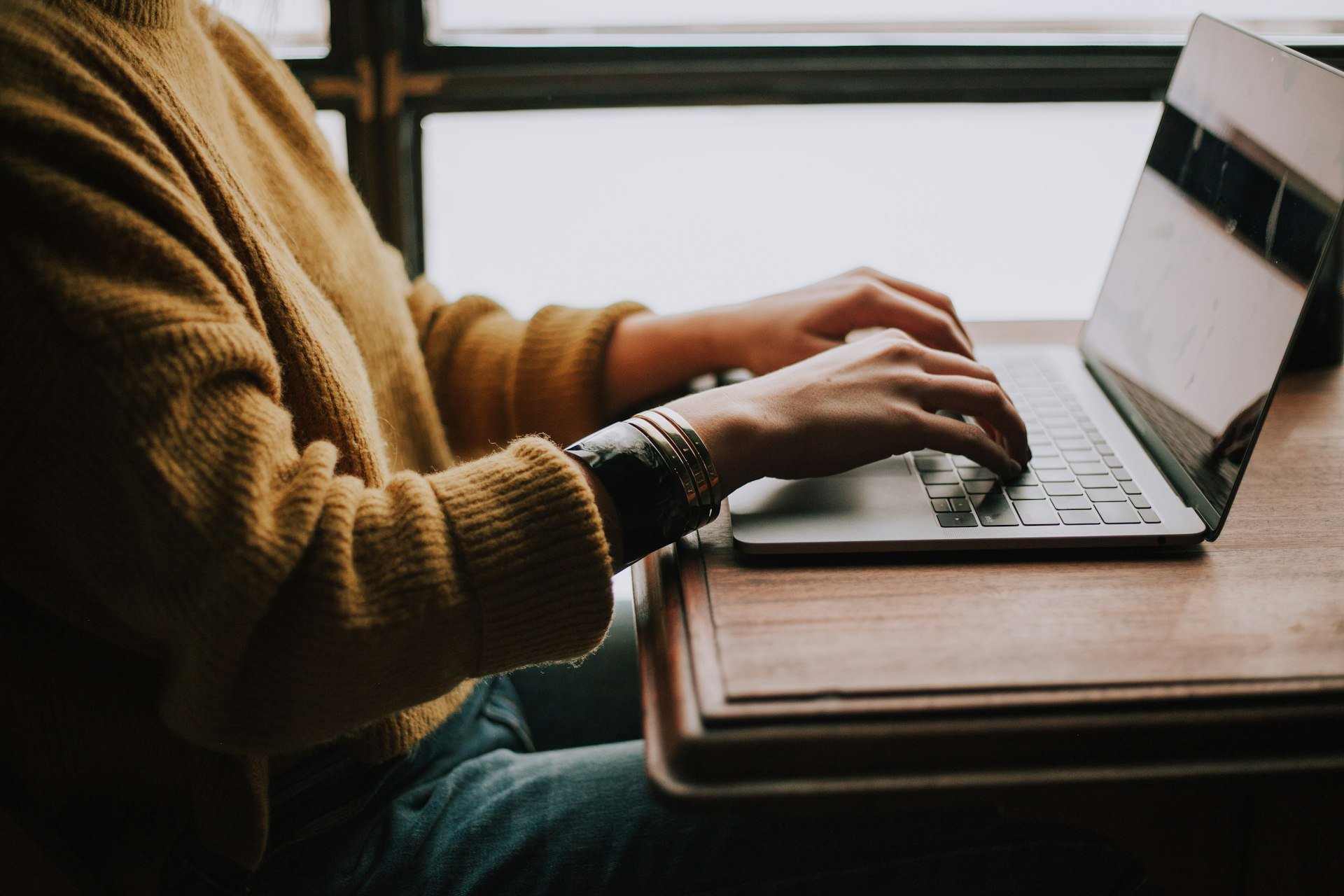 Hands typing on a laptop at a wooden desk beside a window