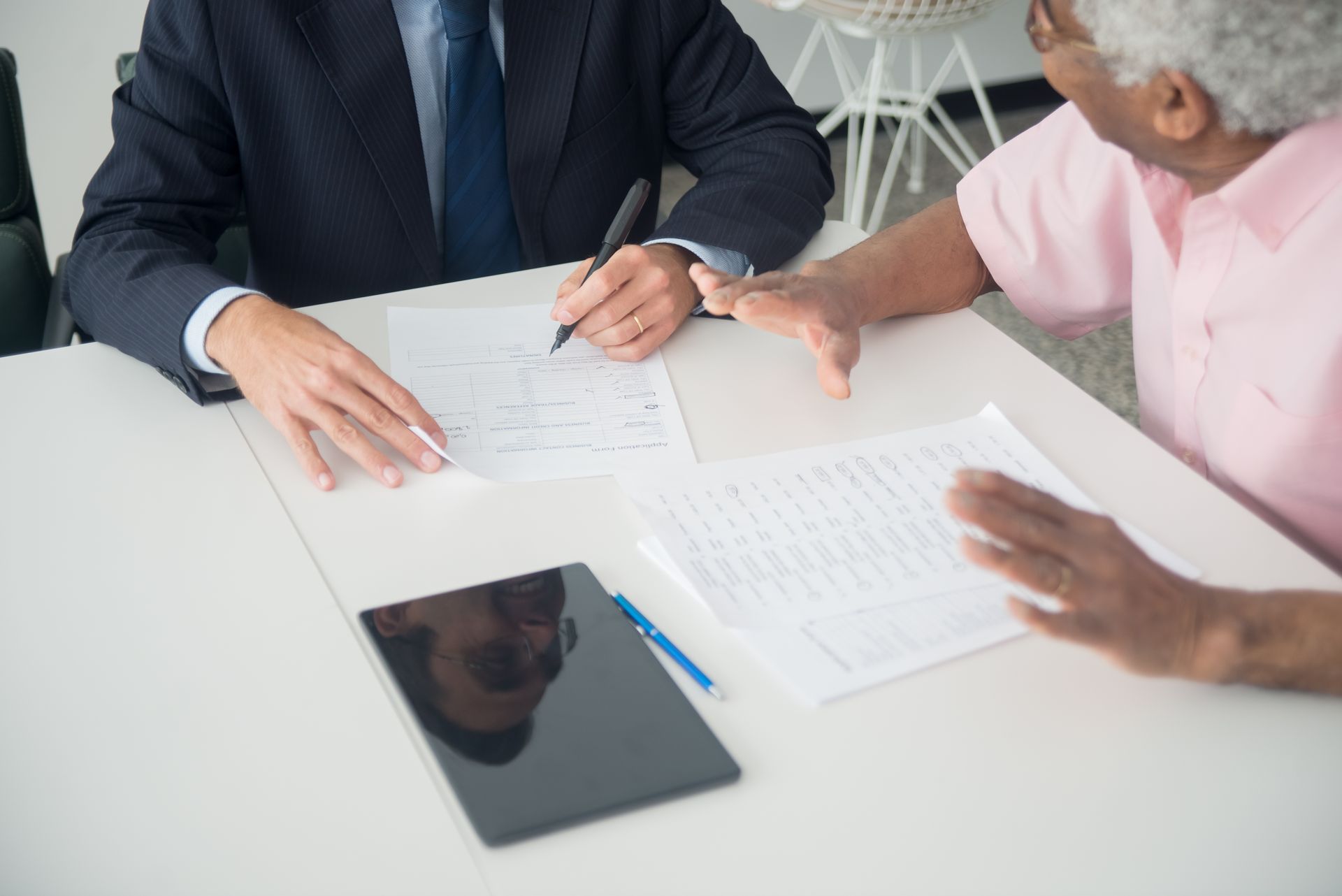Two people at a white table, one holding a pen over printed forms while the other gestures, with a tablet and pen on the table