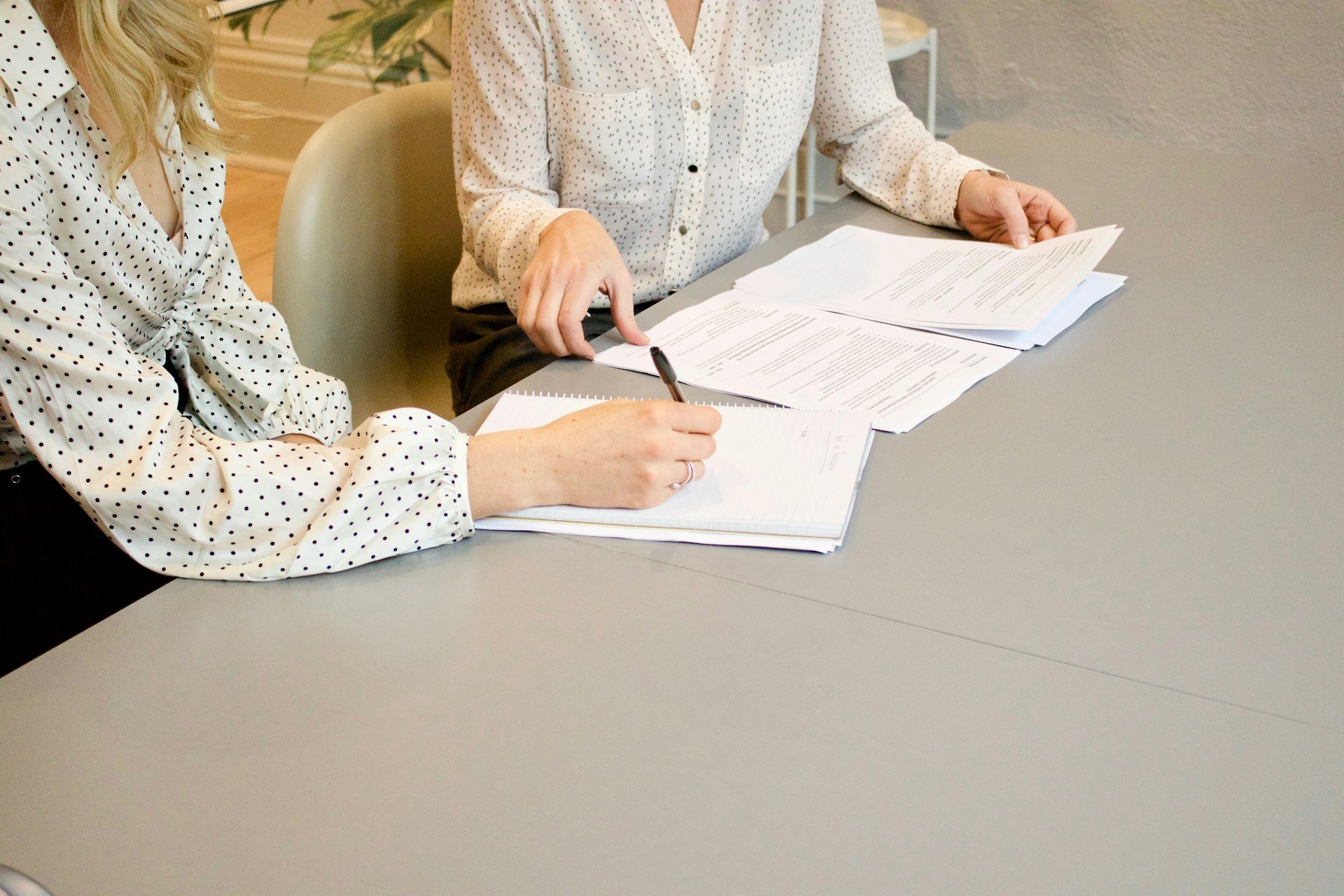 Two people at a grey table, one writing in a notebook while the other turns pages of printed documents