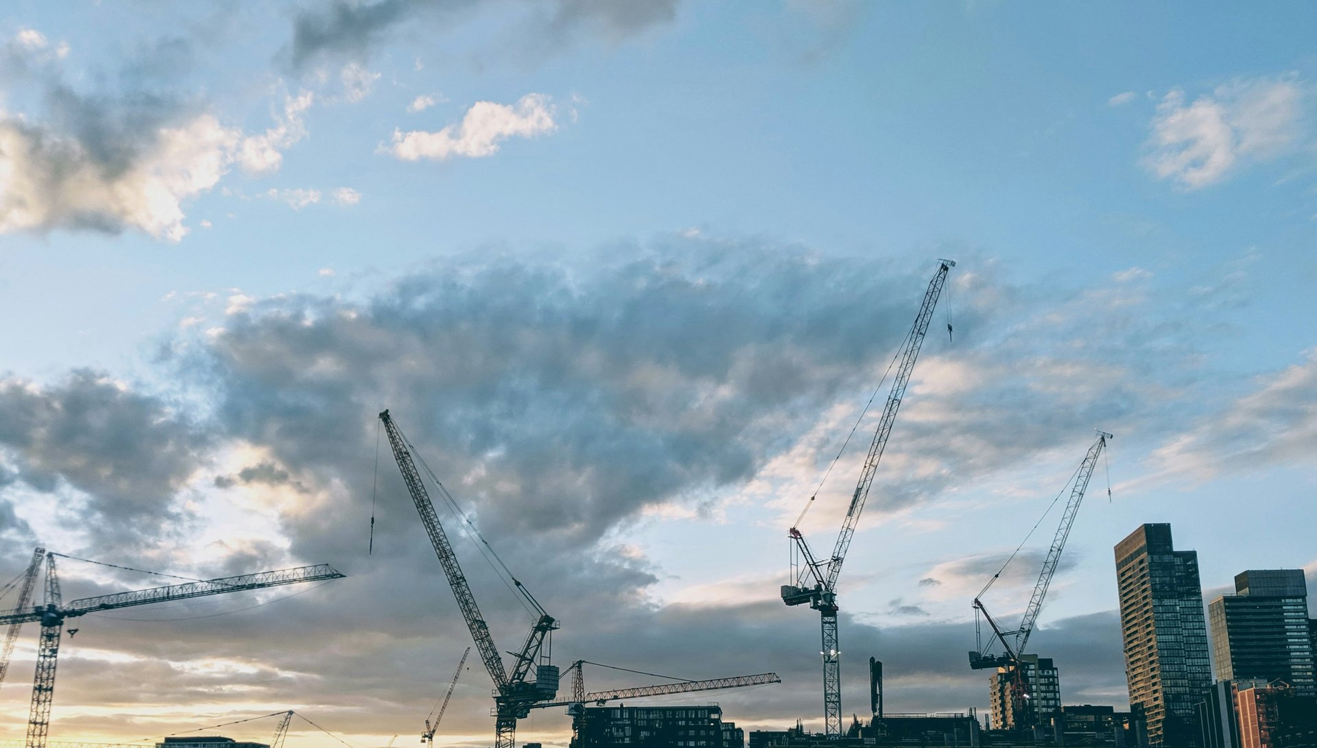 Tower cranes against a cloudy blue sky above city buildings at dusk