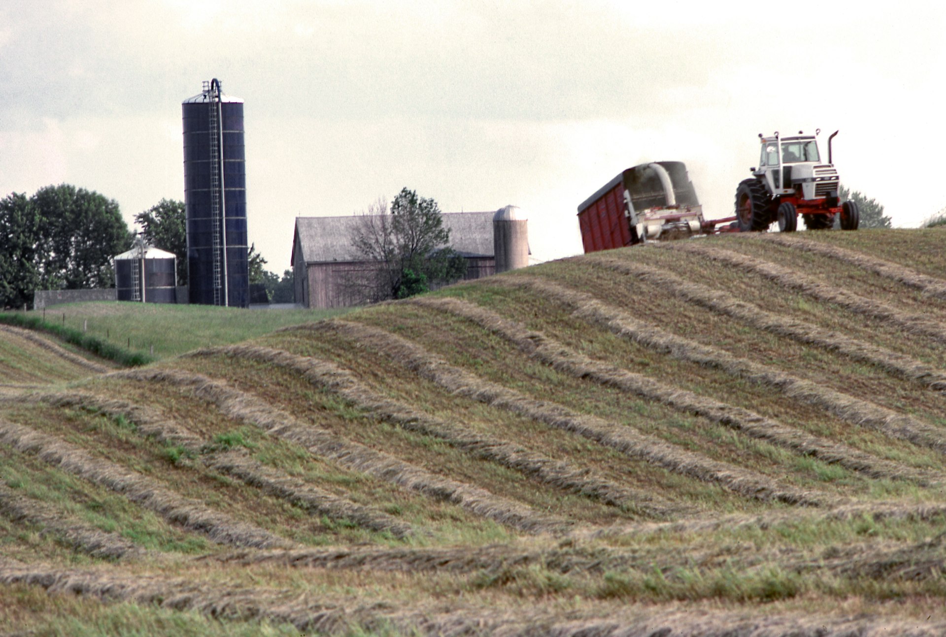 Tractor pulling a red wagon along rows of cut hay on a hillside, with a tall silo and barns behind
