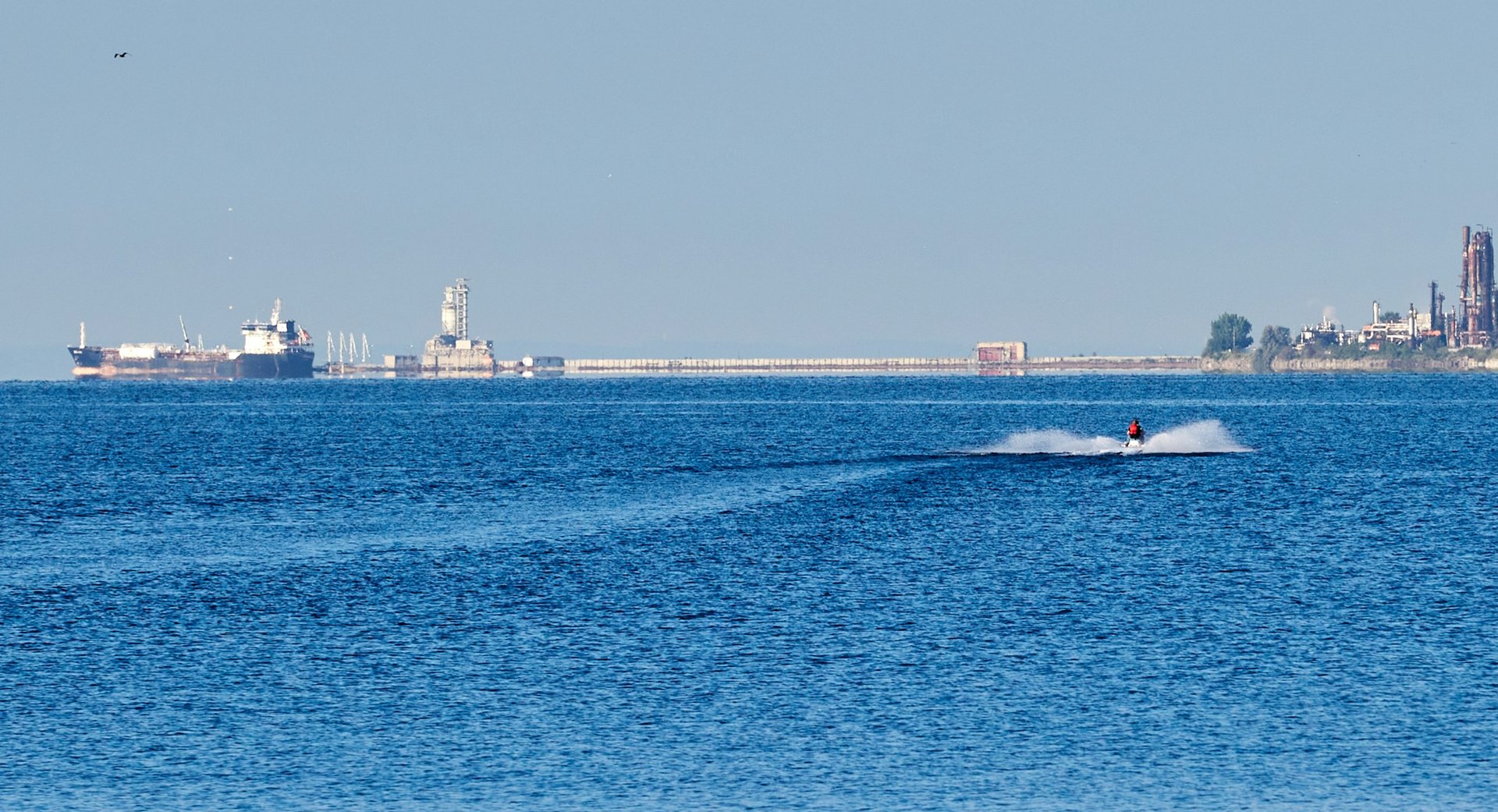 Distant cargo ship and industrial shoreline across open blue water, with a personal watercraft throwing spray