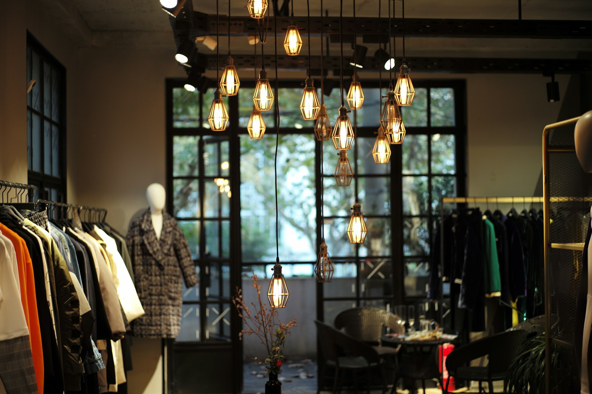 Clothing racks and a mannequin in a boutique with hanging filament lamps and tall windows