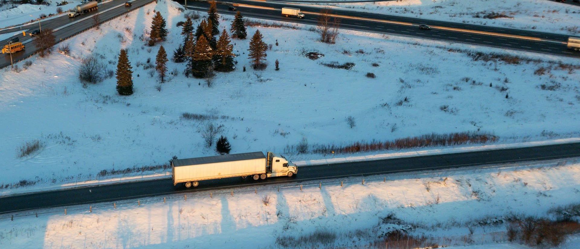 Aerial view of a white transport truck and trailer driving along a snowy highway beside an interchange