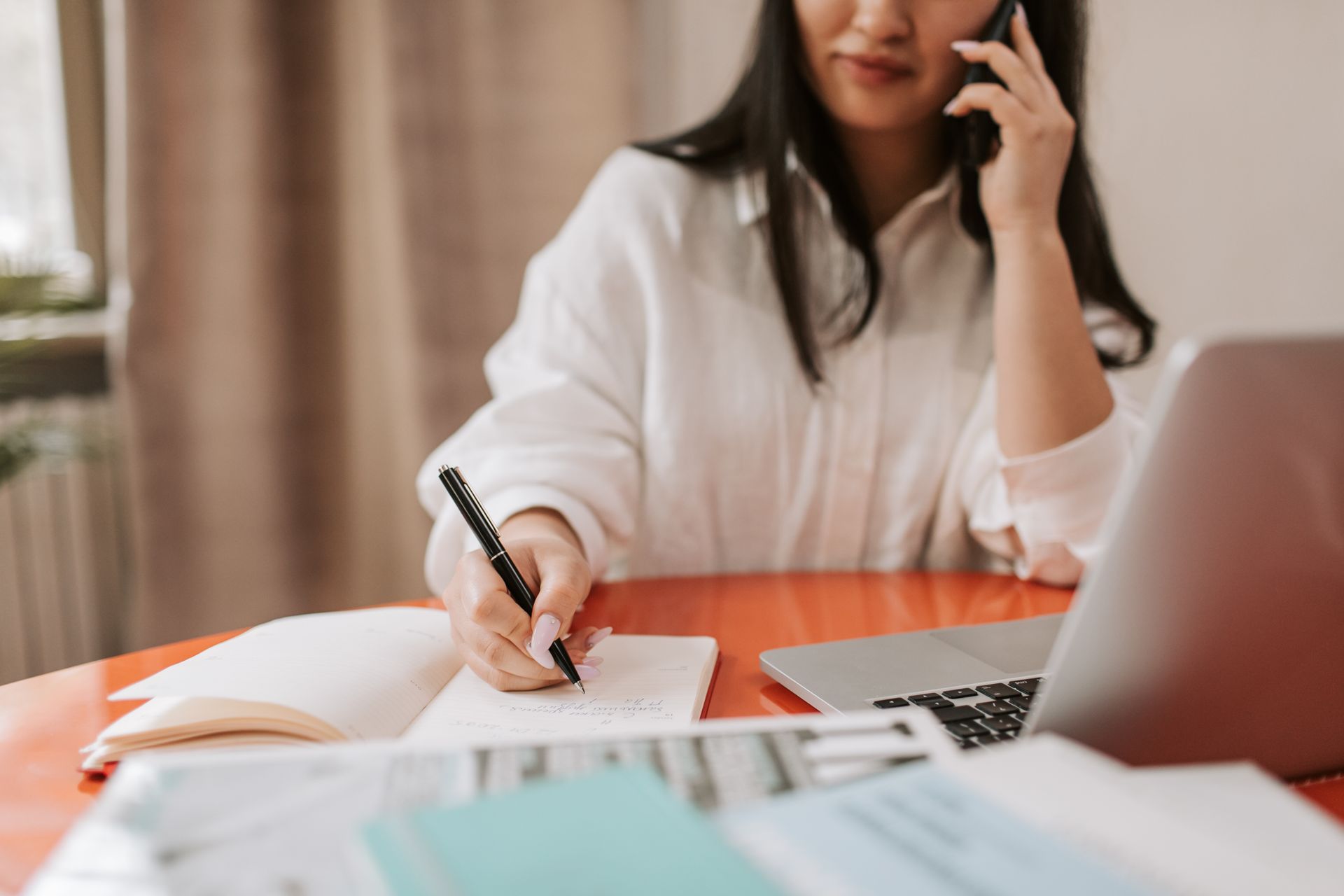 Person on a phone call writing in a notebook at a table with a laptop and papers