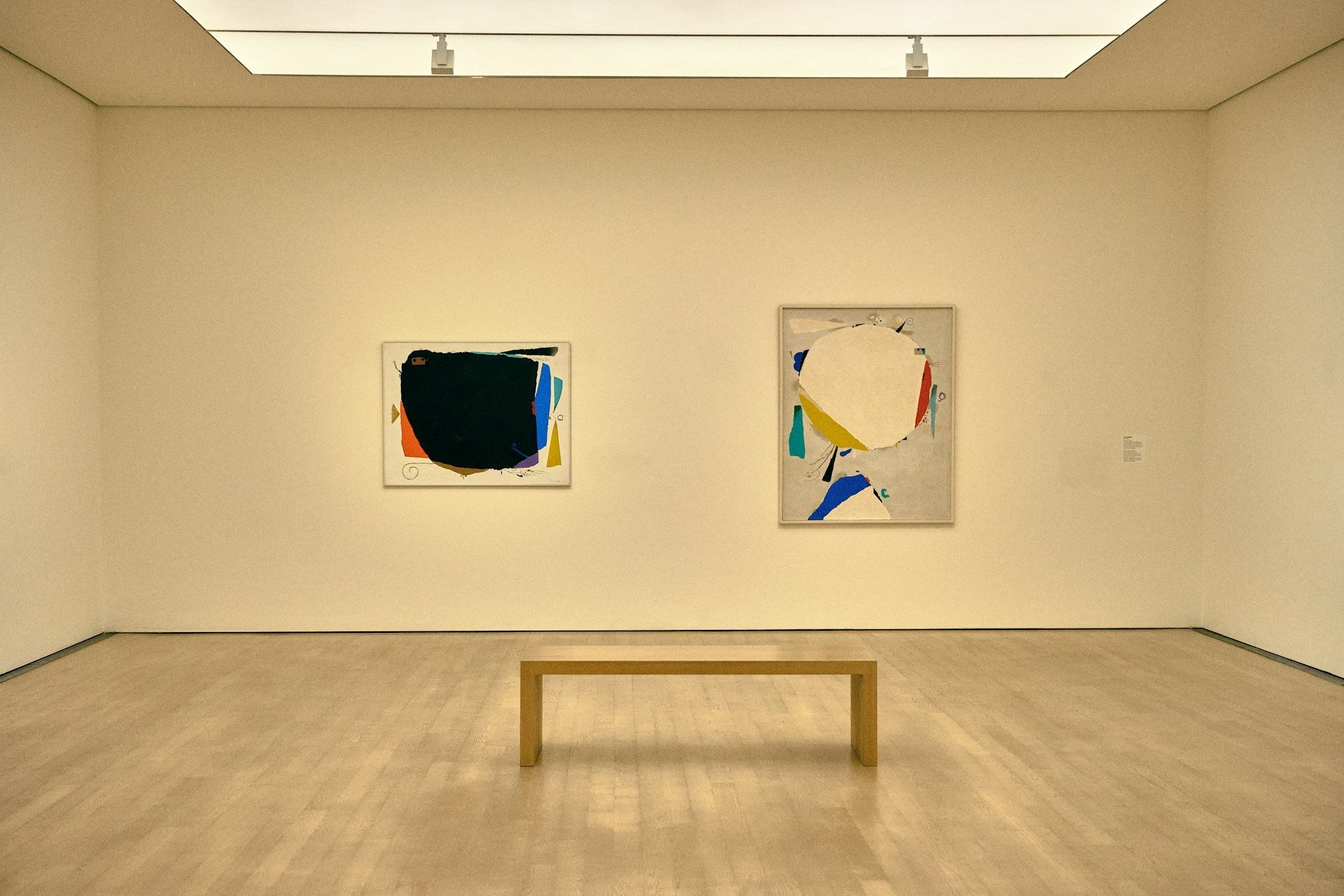 Gallery room with two large abstract paintings on a cream wall and a wooden bench