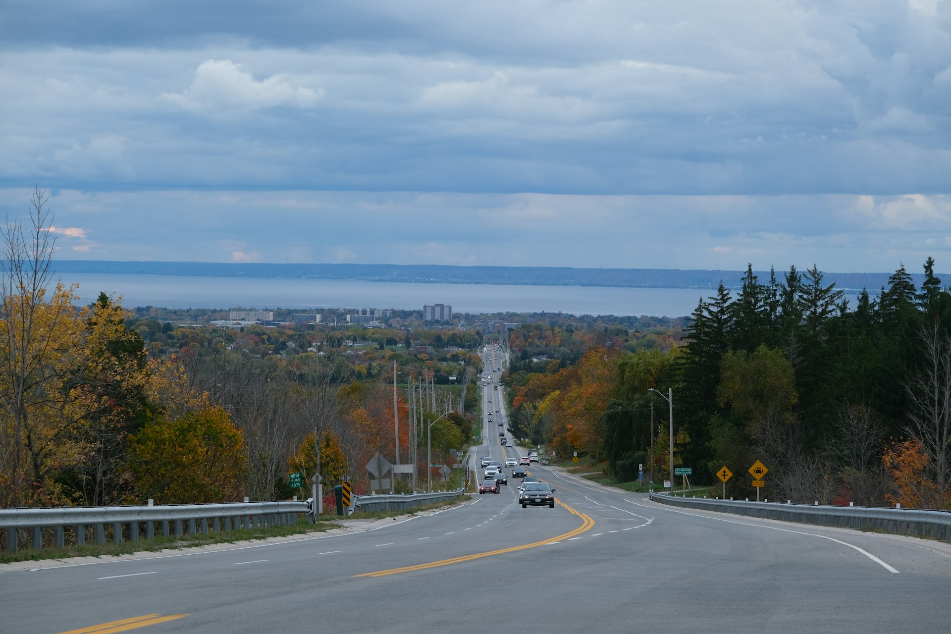 Cars on a long road running down a hillside in autumn toward a distant stretch of water