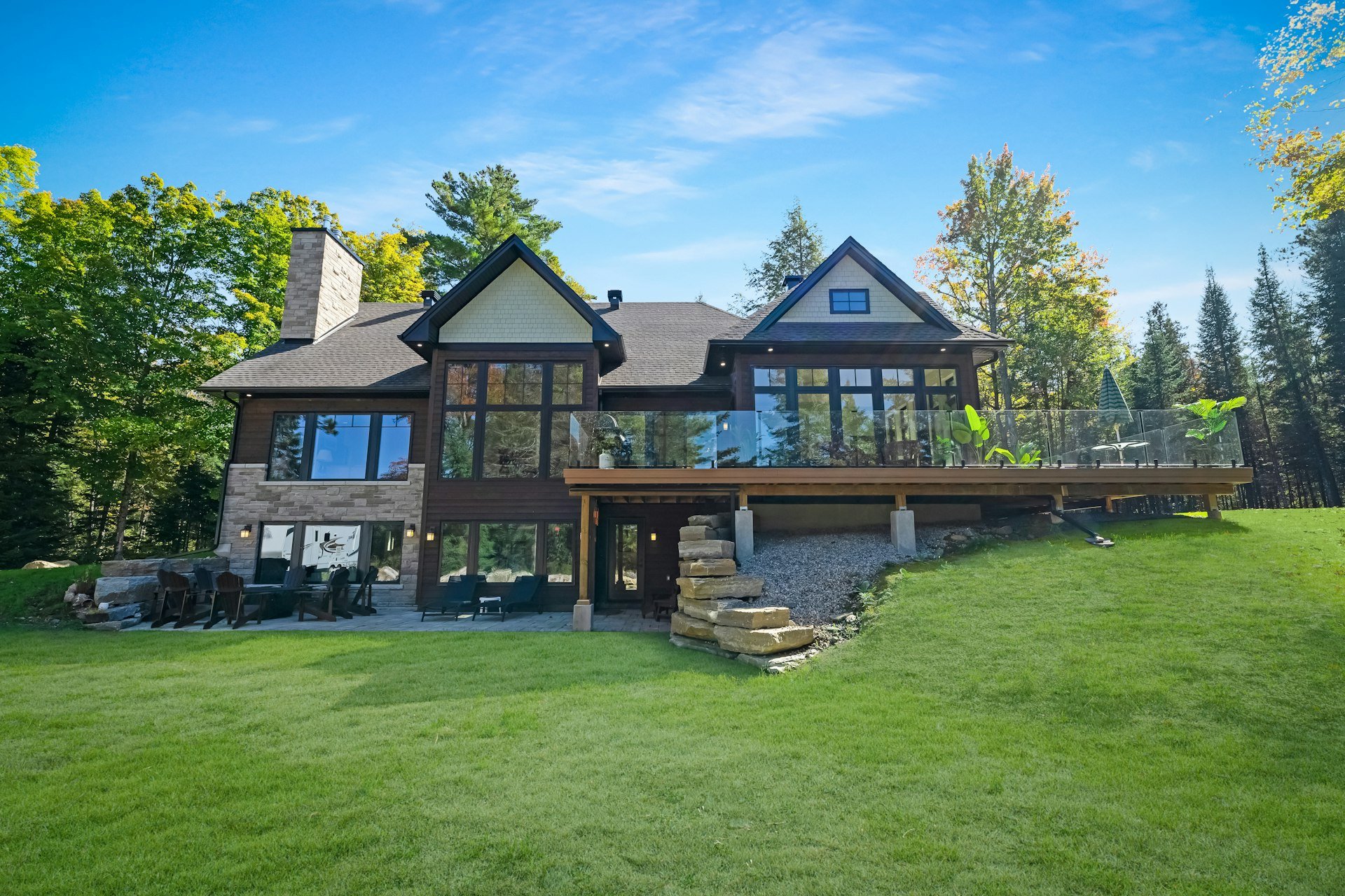 Large stone and timber house with a glass-railed deck above a wide lawn, surrounded by trees