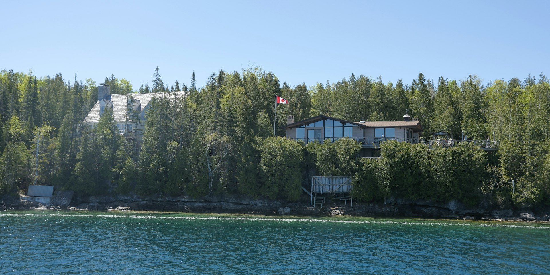 Lakeside homes among evergreen trees above a rocky shoreline, with a Canadian flag on a pole
