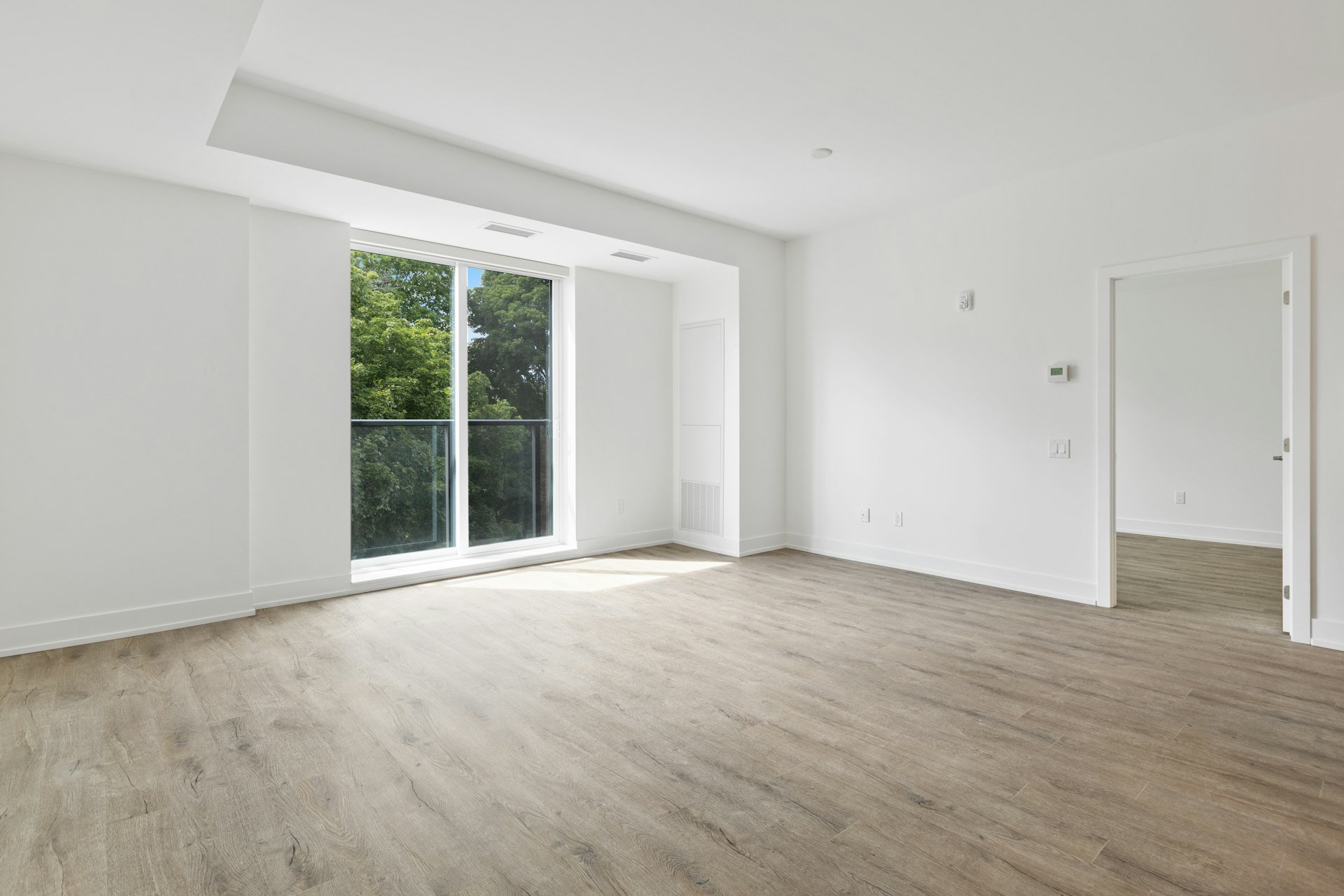 Empty bright apartment living room with a large balcony door, wood-look flooring and an open doorway