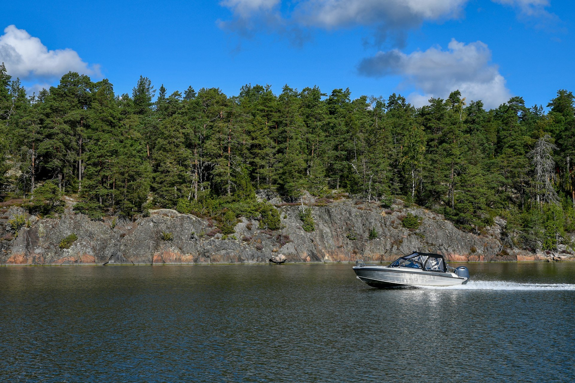 Aluminum cabin boat with a canopy and outboard motor cruising past a rocky, tree-lined shore